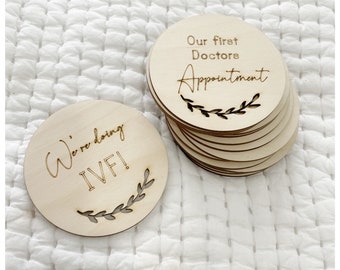 Op de afbeelding: Twee ronde houten schijven met gegraveerde tekst. De eerste schijf zegt "We're doing IVF!" met een bladontwerp. De tweede schijf zegt "Our first Doctors Appointment" met een bladontwerp.