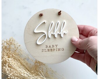 May include: A round wooden sign with the text "Shhh Baby Sleeping" in brown lettering. The sign is white with a white "Shhh" in a cursive font.