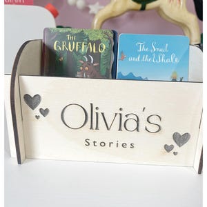 Peut inclure: Un porte-livres en bois avec les mots "Olivia's Stories" gravés sur le devant. Le porte-livres a deux découpes en forme de cœur de chaque côté et contient deux livres pour enfants, "The Gruffalo" et "The Snail and the Whale."