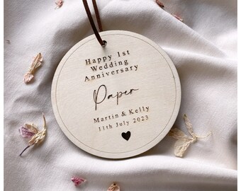 May include: A round wooden ornament with the text "Happy 1st Wedding Anniversary" and "Paper" engraved on it. The names "Martin & Kelly" and the date "11th July 2023" are also engraved. A brown leather cord is attached for hanging.