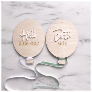 Può includere: Due targhe di legno a forma di palloncino con testo bianco. Una dice "Hello little one" e l'altra "Earth side". Ognuna ha un nastro attaccato. Lo sfondo è una superficie di marmo bianco.