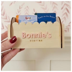 May include: A light-coloured wooden book holder with a scalloped top edge. The holder has the name "Bonnie's stories" in pink letters. Several children's books are inside the holder.