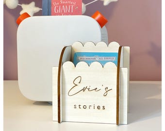 Könnte beinhalten: Eine weiße Nachttischlampe mit orangefarbenen Akzenten und einem Buchhalter aus Holz. Der Buchhalter ist mit "Evie's stories" graviert und enthält zwei Kinderbücher. Im Hintergrund hängt eine dekorative Girlande.