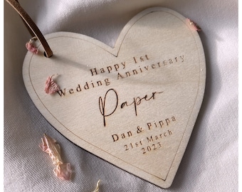May include: A heart-shaped wooden ornament featuring the engraved text Happy 1st Wedding Anniversary, Paper, Dan & Pippa, and 21st March 2023. The light-colored wood piece includes a brown leather cord for hanging and is decorated with small pink flower petals.