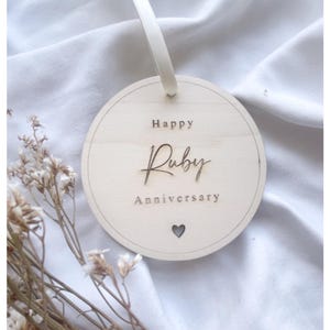 Puede incluir: Un adorno redondo de madera con el texto "Happy Ruby Anniversary" y un pequeño corazón recortado. El adorno está colgado de una cinta blanca.