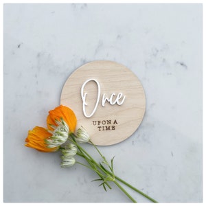 Op de afbeelding: Een rond houten bord met de woorden "Once Upon A Time" in bruine tekst. Het woord "Once" is in witte cursieve letters boven de bruine tekst geschreven. Het bord ligt op een wit marmeren oppervlak met oranje bloemen op de voorgrond.
