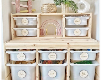 May include: A wooden toy storage unit with white plastic bins labeled with wooden circles. The labels read "Wooden Toys", "Letters and Numbers", "Play Dough", "Natural Resources", "Crafts", "Animals", "Small World", and "People". The unit has a top shelf with a rainbow-colored abacus and a woven basket.