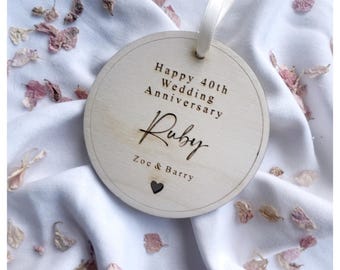May include: A round wooden sign with a white ribbon hanging from the top. The sign says "Happy 40th Wedding Anniversary Ruby Zoc & Barry" with a small heart cutout in the bottom right corner.