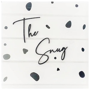 Peut inclure: Une pancarte en bois blanc avec une inscription en écriture cursive noire qui dit "The Snug" avec des points noirs éparpillés sur la surface.