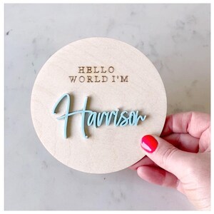 May include: A round wooden sign with the text "Hello World I'm Harrison" in brown lettering. The name Harrison is in light blue raised lettering.