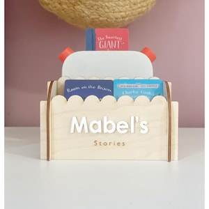 Peut inclure: Un organiseur de livres en bois clair avec le nom "Mabel's Stories" en lettres blanches. Il contient des livres pour enfants, dont "Room on the Broom" et "Charlie Cook's Favorite Book". Le dessus a des détails rouges et blancs.