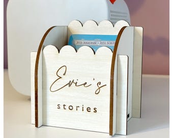 Könnte beinhalten: Ein weißer Holz-Geschichtenhalter mit einer gewellten Oberseite. Der Halter hat die Worte "Evie's Stories" auf der Vorderseite eingraviert.