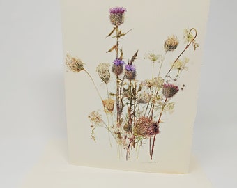 May include: A pressed flower card featuring a bouquet of purple and brown wildflowers, including thistles and Queen Anne's Lace.