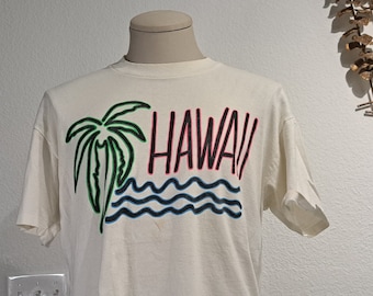 May include: Cream-coloured t-shirt with a colourful graphic of a palm tree, ocean waves, and the word "HAWAII" in red and black. The design is outlined in green and blue. The shirt is displayed on a mannequin.