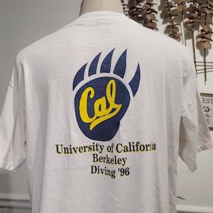 以下が含まれることがあります： 青い熊の足跡と「Cal University of California Berkeley Diving '96」の文字が書かれた白いTシャツ