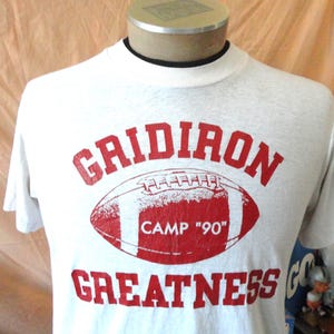 May include: A white short-sleeve t-shirt featuring a red graphic of a football in the center. The text "GRIDIRON" is printed in red above the football, and "GREATNESS" is printed in red below. The football contains the text "CAMP '90".