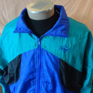 Könnte beinhalten: Eine Vintage-Windjacke in Türkis, Blau und Schwarz. Die Jacke hat einen durchgehenden Reißverschluss, einen Stehkragen und ein kleines Logo auf der Brust. Das Colorblock-Design zeigt eine Mischung aus einfarbigen und kontrastierenden Einsätzen.