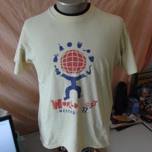 May include: Cream-coloured t-shirt with a graphic of a globe with a red grid, blue figure, and musical instrument icons. The text "World Fest Westpo 97" is printed below the graphic in red and blue.