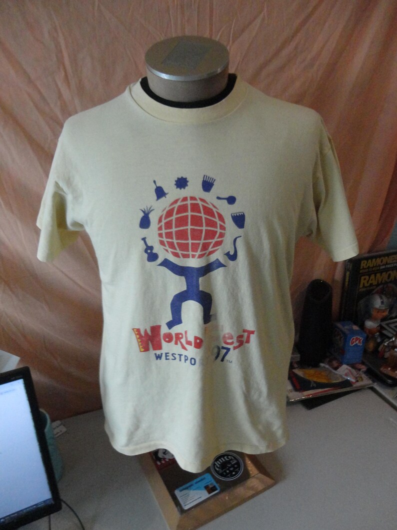 May include: Cream-coloured t-shirt with a graphic of a globe with a red grid, blue figure, and musical instrument icons. The text "World Fest Westpo 97" is printed below the graphic in red and blue.