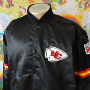 May include: Black satin jacket featuring the Kansas City Chiefs logo on the chest and NFL patch on the sleeve. The jacket has red and yellow stripes on the sleeve cuffs. The collar and button-up front are black.