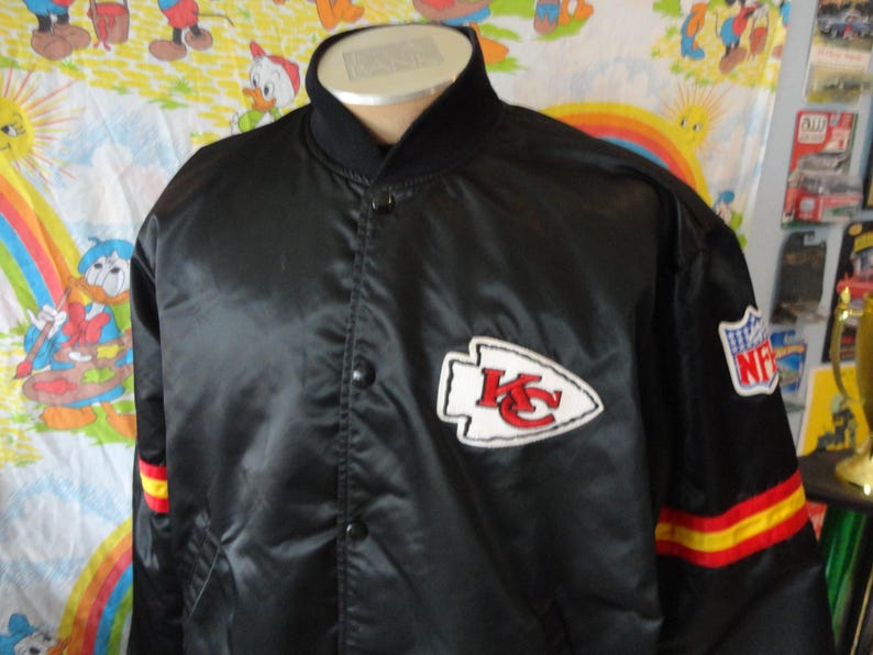 May include: Black satin jacket featuring the Kansas City Chiefs logo on the chest and NFL patch on the sleeve. The jacket has red and yellow stripes on the sleeve cuffs. The collar and button-up front are black.
