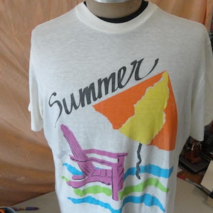 May include: A white short-sleeve t-shirt featuring the word Summer in black script text. Below the text is a graphic of a purple lounge chair under an orange and yellow umbrella, with blue and green wavy lines representing water at the bottom.