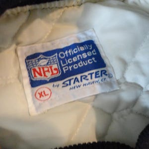 May include: Close-up of a white and quilted jacket lining with a blue and white NFL Officially Licensed Product tag. The tag includes the NFL logo and the Starter brand name, with an XL size indicator.
