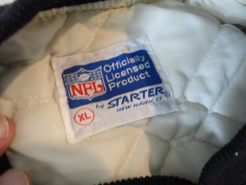 May include: Close-up of a white and quilted jacket lining with a blue and white NFL Officially Licensed Product tag. The tag includes the NFL logo and the Starter brand name, with an XL size indicator.