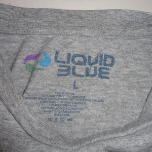 May include: Close-up of a gray t-shirt collar with the "LIQUID BLUE" logo in blue. The logo includes a colorful design. The size "L" is visible. The shirt's label provides care instructions.