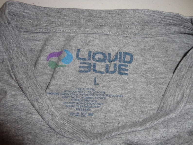 May include: Close-up of a gray t-shirt collar with the "LIQUID BLUE" logo in blue. The logo includes a colorful design. The size "L" is visible. The shirt's label provides care instructions.