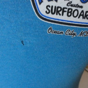May include: Close-up of a light blue t-shirt with a graphic. The graphic reads "Custom Surfboards" in white and blue, with "Ocean City, MD" below in script. The shirt is made of a soft material.
