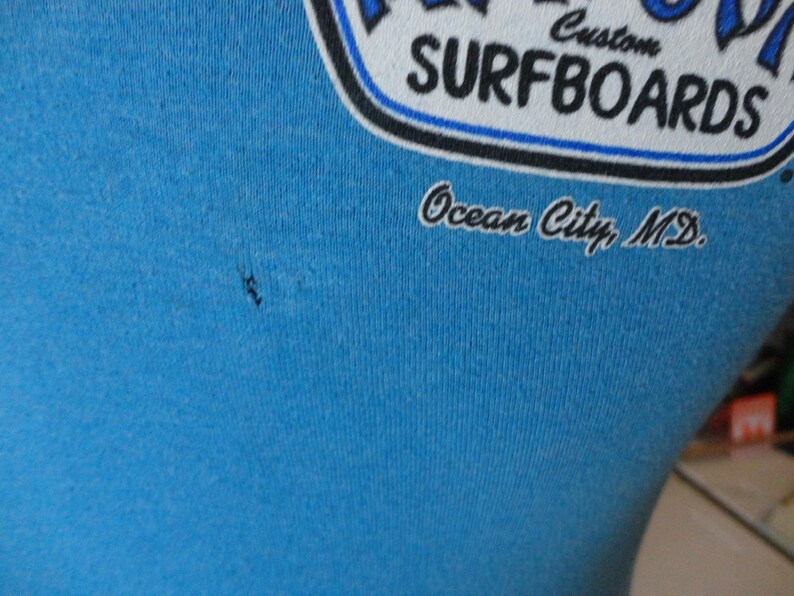 May include: Close-up of a light blue t-shirt with a graphic. The graphic reads "Custom Surfboards" in white and blue, with "Ocean City, MD" below in script. The shirt is made of a soft material.