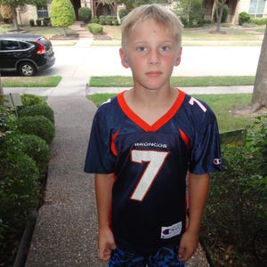 May include: A navy blue short-sleeved sports jersey featuring the word BRONCOS in white lettering above a large white number 7 with orange trim. The V-neck collar is orange and navy blue, and a small white Champion logo is on the left sleeve.