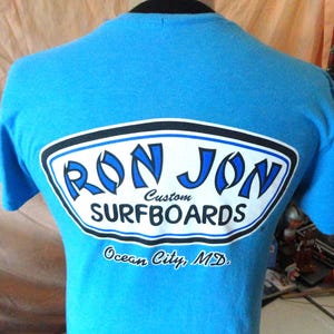 May include: A turquoise t-shirt featuring a white and blue logo that says "RON JON Custom SURFBOARDS". The text "Ocean City, MD" is printed below the logo. The shirt is displayed on a mannequin. The logo has a black border.