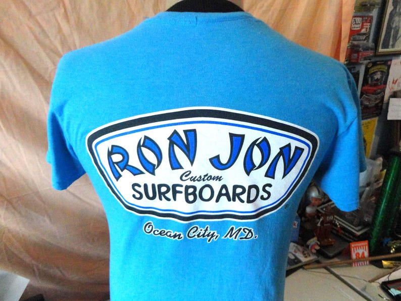 May include: A turquoise t-shirt featuring a white and blue logo that says "RON JON Custom SURFBOARDS". The text "Ocean City, MD" is printed below the logo. The shirt is displayed on a mannequin. The logo has a black border.