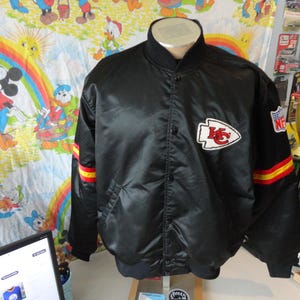 May include: Black satin jacket with the Kansas City Chiefs logo on the chest. The jacket has red and yellow stripes on the sleeves and an NFL patch. The jacket is button-down and has a ribbed collar.