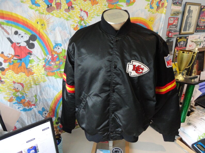 May include: Black satin jacket with the Kansas City Chiefs logo on the chest. The jacket has red and yellow stripes on the sleeves and an NFL patch. The jacket is button-down and has a ribbed collar.