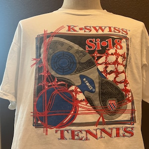 May include: White t-shirt with a graphic of a tennis shoe and a tennis ball. The graphic is in red, blue, and black. The text "K-Swiss Si-18 Tennis" is printed on the shirt.
