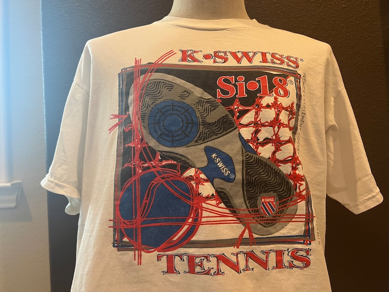 May include: White t-shirt with a graphic of a tennis shoe and a tennis ball. The graphic is in red, blue, and black. The text "K-Swiss Si-18 Tennis" is printed on the shirt.