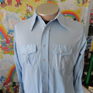 Puede incluir: Camisa abotonada azul claro con dos bolsillos en el pecho. La camisa de manga larga tiene cuello puntiagudo y se exhibe en un maniquí. La camisa es de tejido liso.