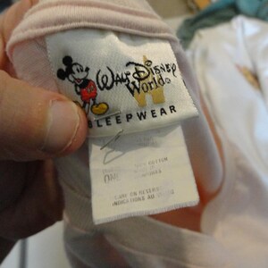 May include: A light pink sleepwear garment with a tag featuring the Walt Disney World logo and a graphic of Mickey Mouse. The tag also includes the word "SLEEPWEAR". The garment is made of 100% cotton.