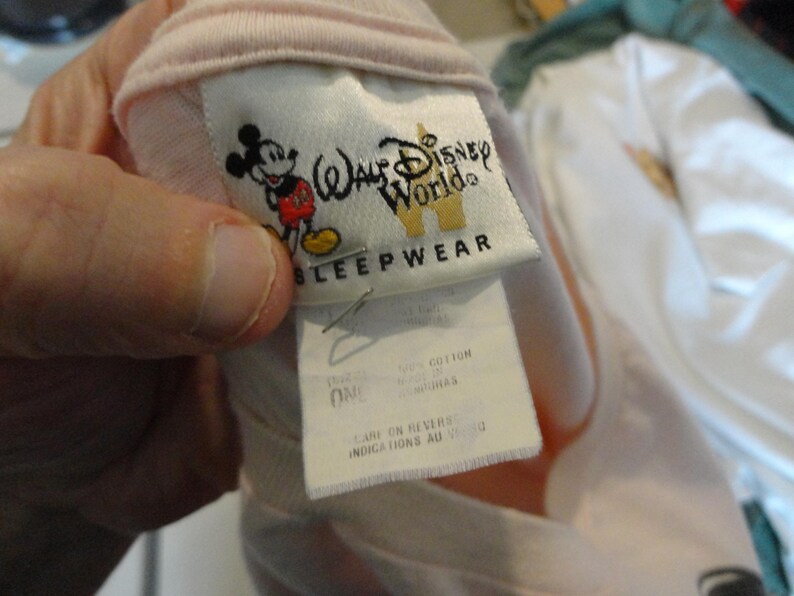May include: A light pink sleepwear garment with a tag featuring the Walt Disney World logo and a graphic of Mickey Mouse. The tag also includes the word "SLEEPWEAR". The garment is made of 100% cotton.
