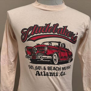 Könnte beinhalten: Ein Vintage-T-Shirt mit langen Ärmeln in Weiß mit einem roten und schwarzen Aufdruck eines roten Cabriolets mit zwei Personen darin. Der Aufdruck enthält auch den Text "Studebaker's 50's, 60's & Beach Music Atlanta, Ga."