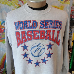 May include: A light grey sweatshirt with the words "WORLD SERIES BASEBALL" in red and blue. Below is a baseball with "BABE RUTH" and blue stars. A vintage sports-themed garment.