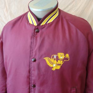 May include: Maroon varsity jacket with a yellow and white striped collar and cuffs. Features a yellow cartoon beaver mascot patch with the letters "M" and "N" on the chest. The jacket has a button-up front and a pocket.