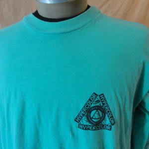 May include: A long-sleeve t-shirt in a bright turquoise color with a crew neckline. The shirt features a black triangular graphic on the lower right side with the word HYPERCOLOR printed three times along the edges of the triangle.