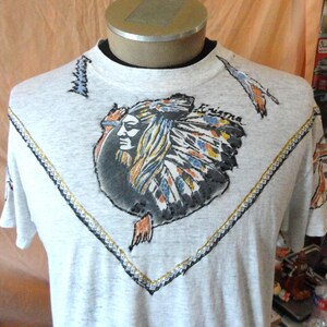 May include: A light gray heathered short-sleeve t-shirt featuring a central graphic of a person wearing a feathered headdress. The design includes a V-shaped border with geometric patterns and the word Enigma printed in black script near the top.