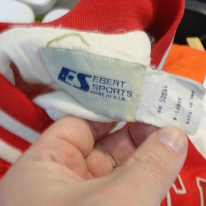 May include: A white and red baseball jacket with white stripes on the sleeves. The jacket has a white label with the text "Ebert Sports Made in U.S.A." and a smaller blue label with the letters "ES". The label also has the text "MN 52894 X-Large Made in Taiwan."