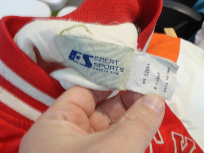 May include: A white and red baseball jacket with white stripes on the sleeves. The jacket has a white label with the text "Ebert Sports Made in U.S.A." and a smaller blue label with the letters "ES". The label also has the text "MN 52894 X-Large Made in Taiwan."