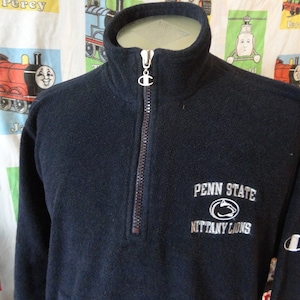 Puede incluir: Una sudadera de vellón negra Champion con cierre de cremallera de media cremallera. La sudadera tiene un logotipo bordado blanco en el pecho izquierdo y un logotipo bordado blanco de Penn State Nittany Lions en la parte delantera.